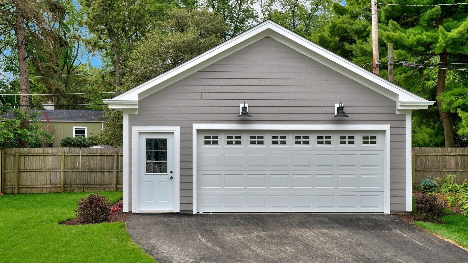 Modern gray garage with white trim, white door, and driveway surrounded by green lawn and trees