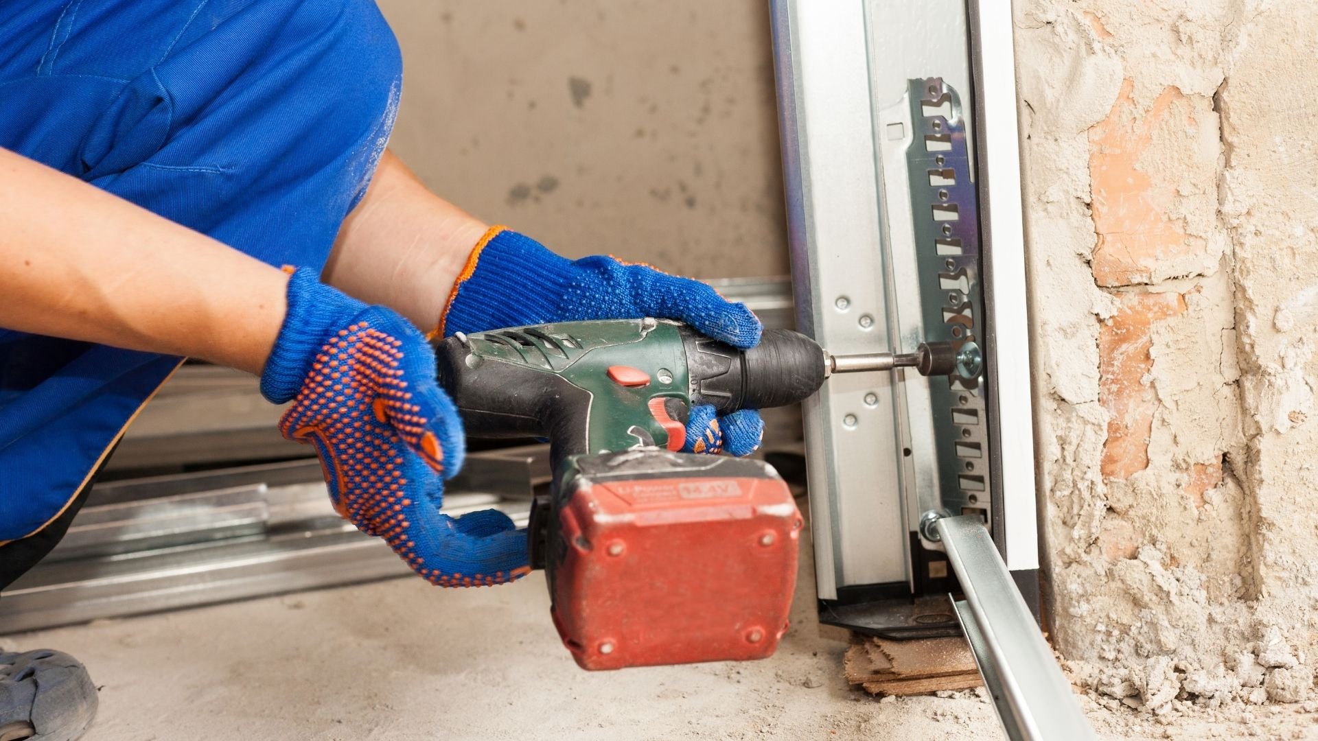 Worker in blue gloves using power drill on metal stud frame against brick wall